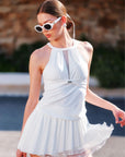 Woman wearing a white pleated dress and sunglasses by a poolside.
