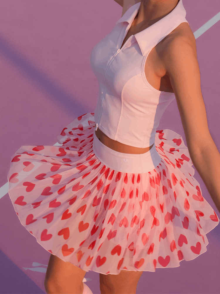Model wearing the Mesh Tennis Skirt - Pink Heart, twirling on a pink tennis court to show the flowing heart-print mesh fabric