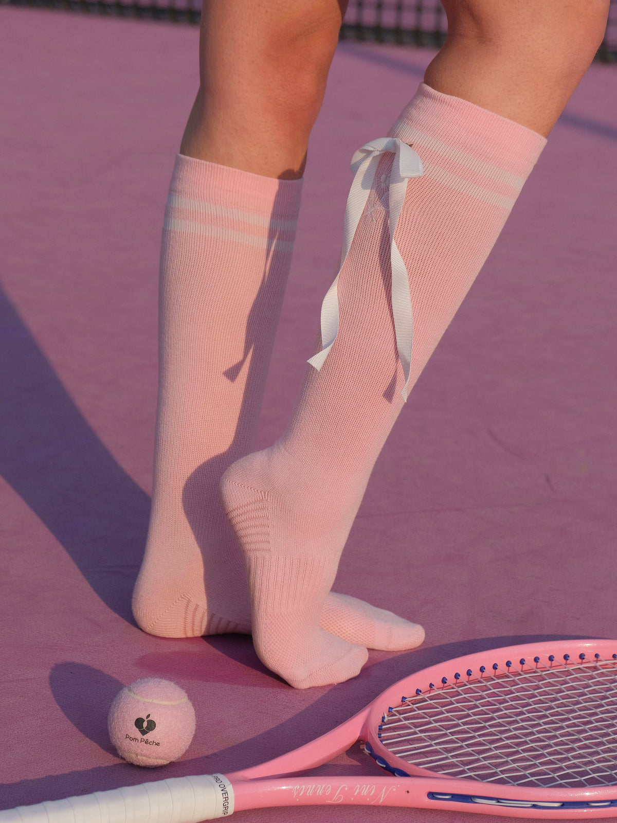 Soft pink knee-high tennis socks with white stripes, bow detail, and logo embroidery.