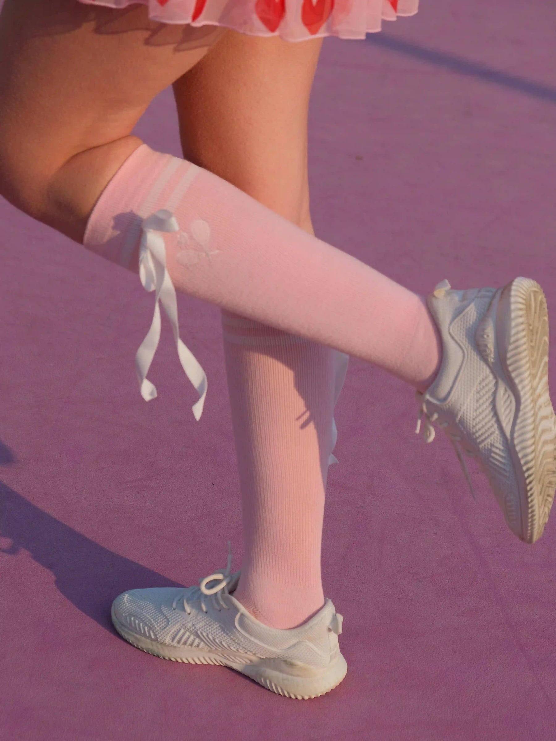 Person wearing pink knee-high socks with white bows and light blue sneakers on a purple floor.