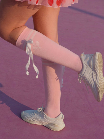 Soft pink knee-high tennis socks with white stripes, bow detail, and logo embroidery.