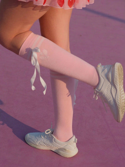 Person wearing pink knee-high socks with white bows and light blue sneakers on a purple floor.