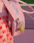 Pink backpack with a heart-patterned dress and tennis ball keychain on a tennis court.