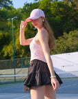 Woman on a tennis court wearing a pink cap and skirt, with trees in the background.