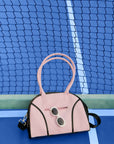 Soft pink dome-shaped tennis bag featuring Pom Pêche logo, styled with black trim and placed on a tennis court.