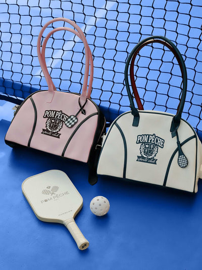 Soft pink dome-shaped tennis bag featuring Pom Pêche logo, styled with black trim and placed on a tennis court.