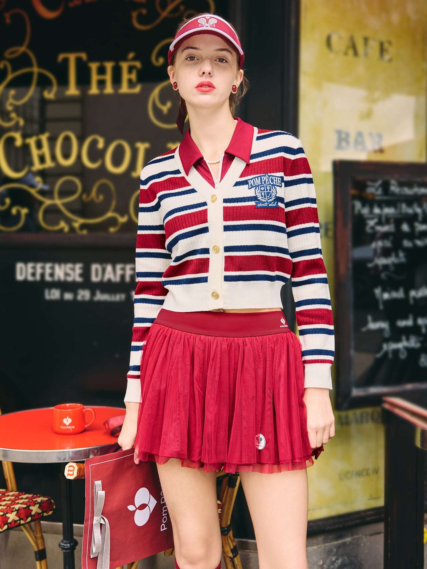 Red, navy, and white striped cardigan with a V-neck, button-down design, featuring an embroidered logo. Styled from court to café.