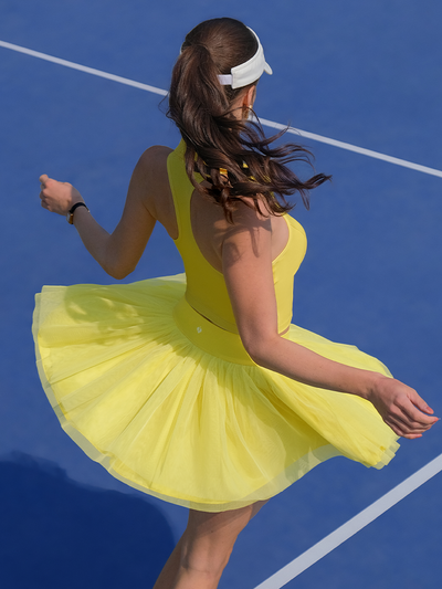 Model wearing a yellow tulle puff tennis skirt spinning on blue court, showing airy layered movement and a high-waisted tennis skirt fit.