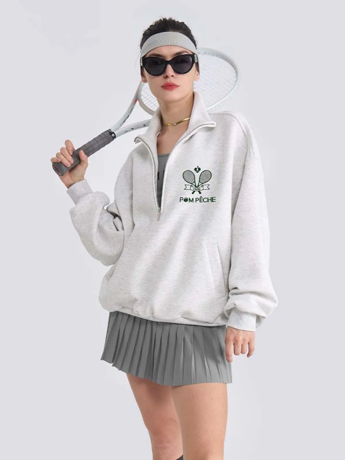 Person wearing a white sweatshirt with a logo and gray pleated skirt, holding a tennis racket.