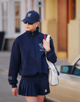Person wearing a navy blue outfit with a cap and sunglasses, holding a white bag.