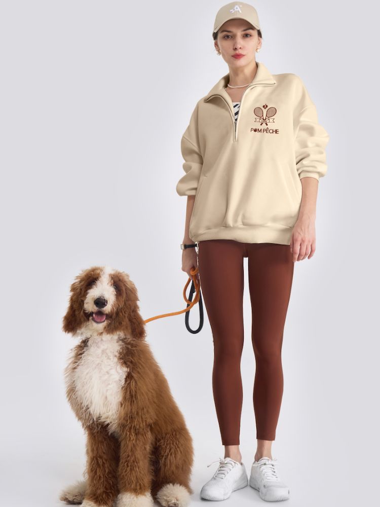 Person wearing a beige hoodie and brown leggings standing next to a dog on a white background