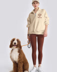 Person wearing a beige hoodie and brown leggings standing next to a dog on a white background