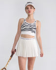 The model is showcasing a white pleated tennis skirt in a clean athletic stance on the court.