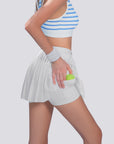 The model is revealing built-in shorts under a white pleated tennis skirt while holding a tennis ball.