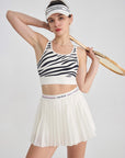 The model is wearing a white pleated tennis skirt while posing confidently with a sporty racket.