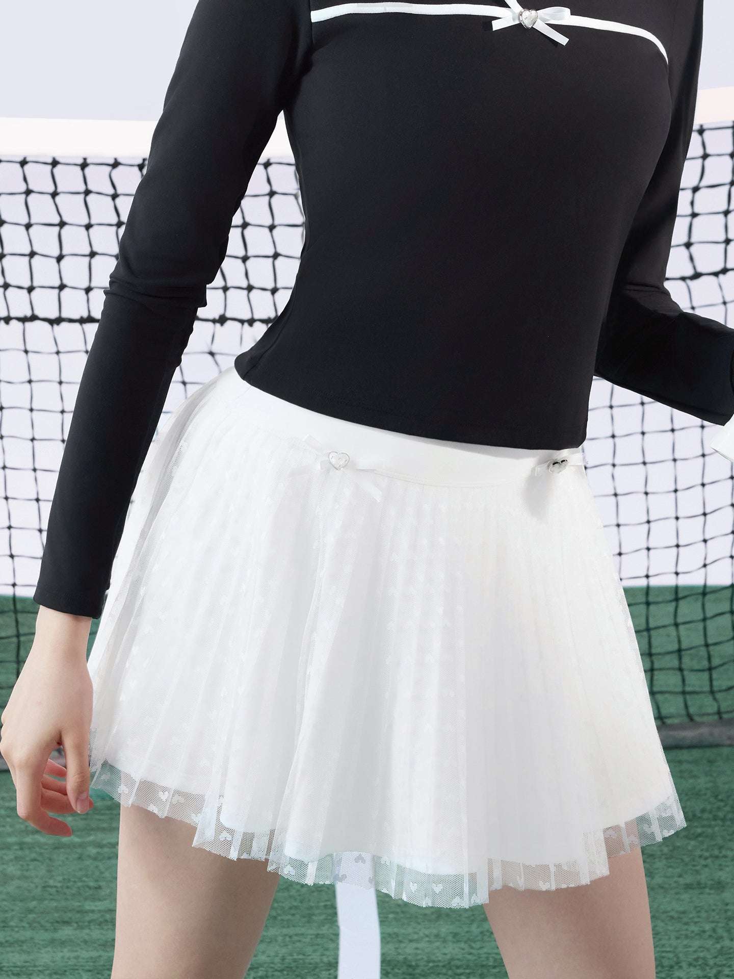 White pleated tennis skirt with soft mesh lace and subtle heart details, offering a lightweight feel, styled on a tennis court. 