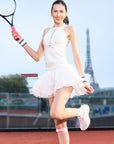 Model wearing white mesh halter tank and petal tiered tennis skirt swinging a racket on a tennis court, showing fluid movement and leg-lengthening effect.