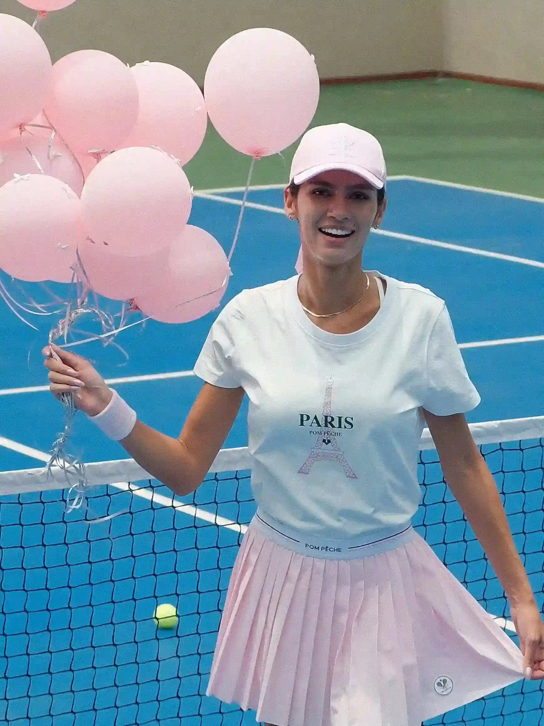 Woman holding pink balloons on a tennis court wearing a 'PARIS' t-shirt.