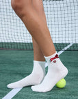 White ankle tennis socks with red bow detail and Pom Pêche logo, styled on the court