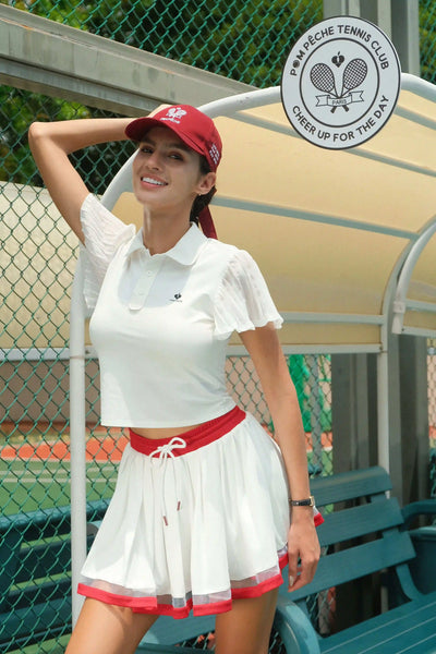 Woman in a white tennis dress with red accents standing on a tennis court, wearing a red cap.