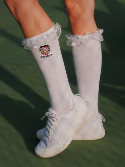 White knee-high socks with ruffles and a logo, worn with white sneakers on a green background.