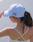 Woman wearing a white cap with text and a white sports bra with thin straps.