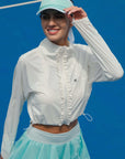 White cropped jacket with ruffled zip-front detail and long sleeves, styled on the tennis court.