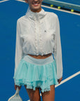 White cropped jacket with ruffled zip-front detail and long sleeves, styled on the tennis court.