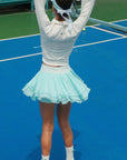 White cropped jacket with ruffled zip-front detail and long sleeves, styled on the tennis court.