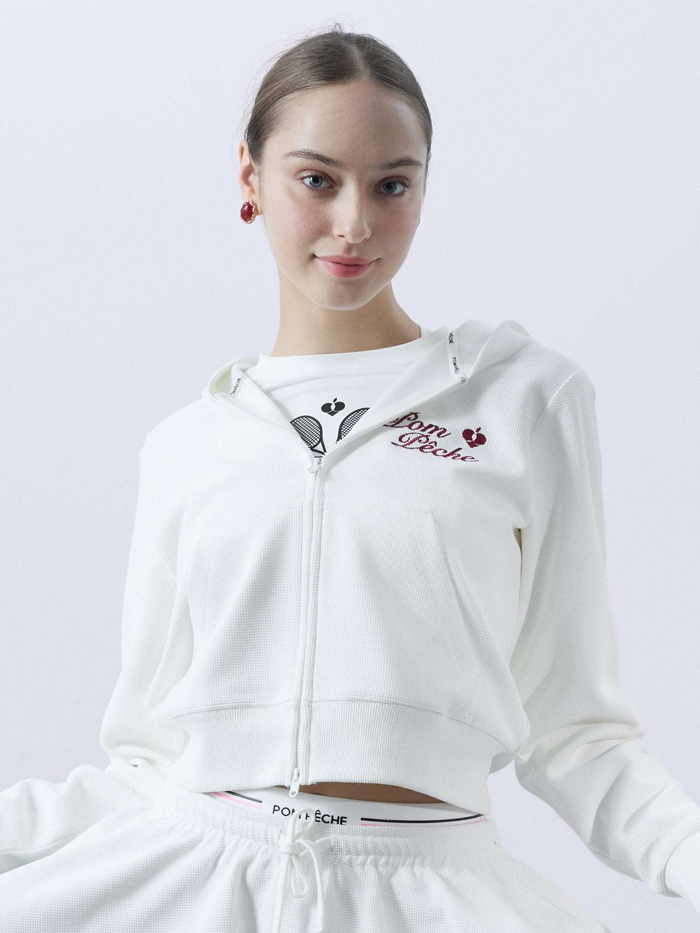 White zip-up hoodie with bow detail on the hood and embroidered logo, perfect for warm-ups or everyday wear.