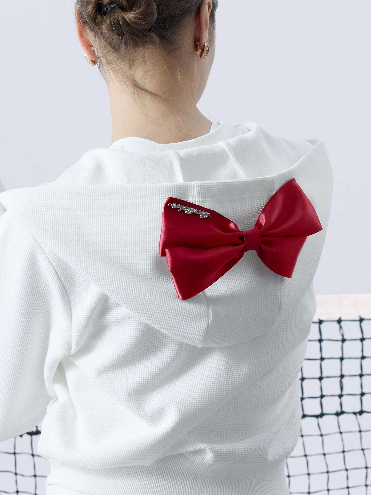 White zip-up hoodie with bow detail on the hood and embroidered logo, perfect for warm-ups or everyday wear.
