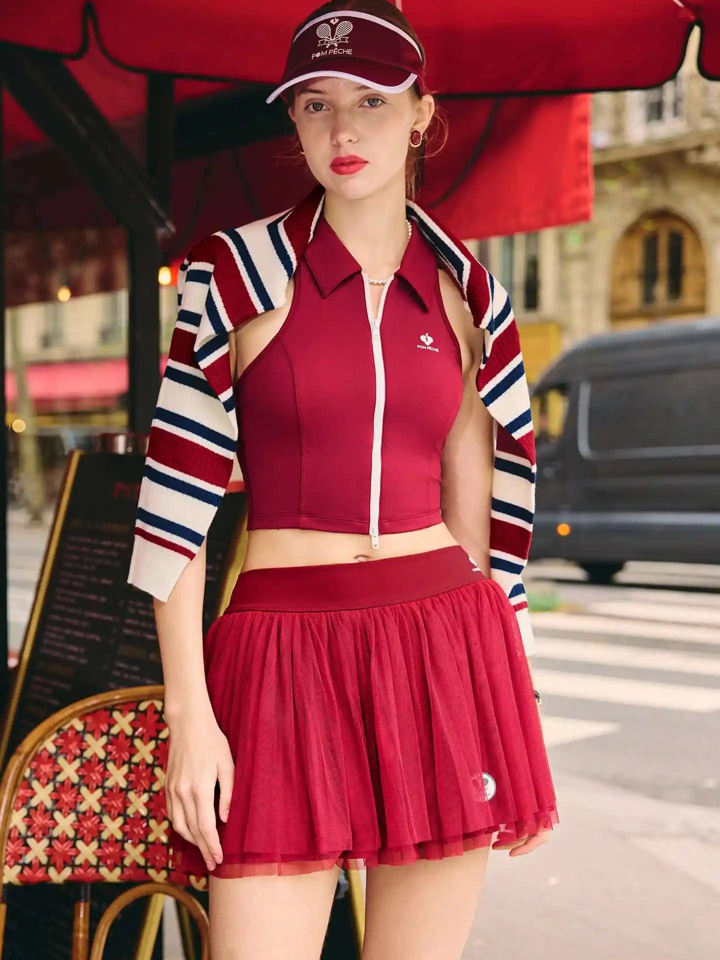 Woman in a red outfit with a striped jacket standing under a red umbrella in an urban setting.