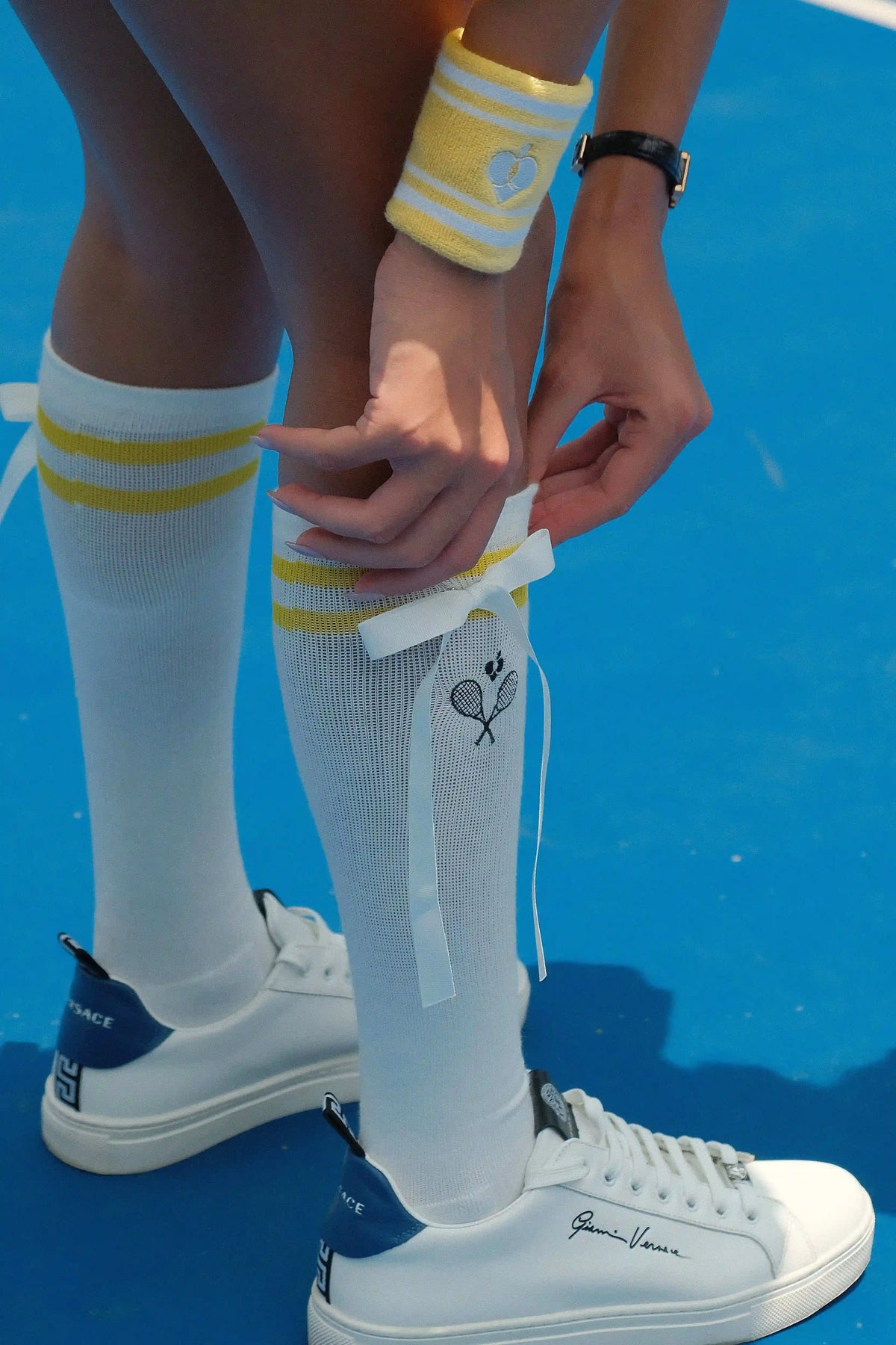 Person wearing white sneakers with a logo, white socks with yellow stripes, and a yellow wristband on a blue background.