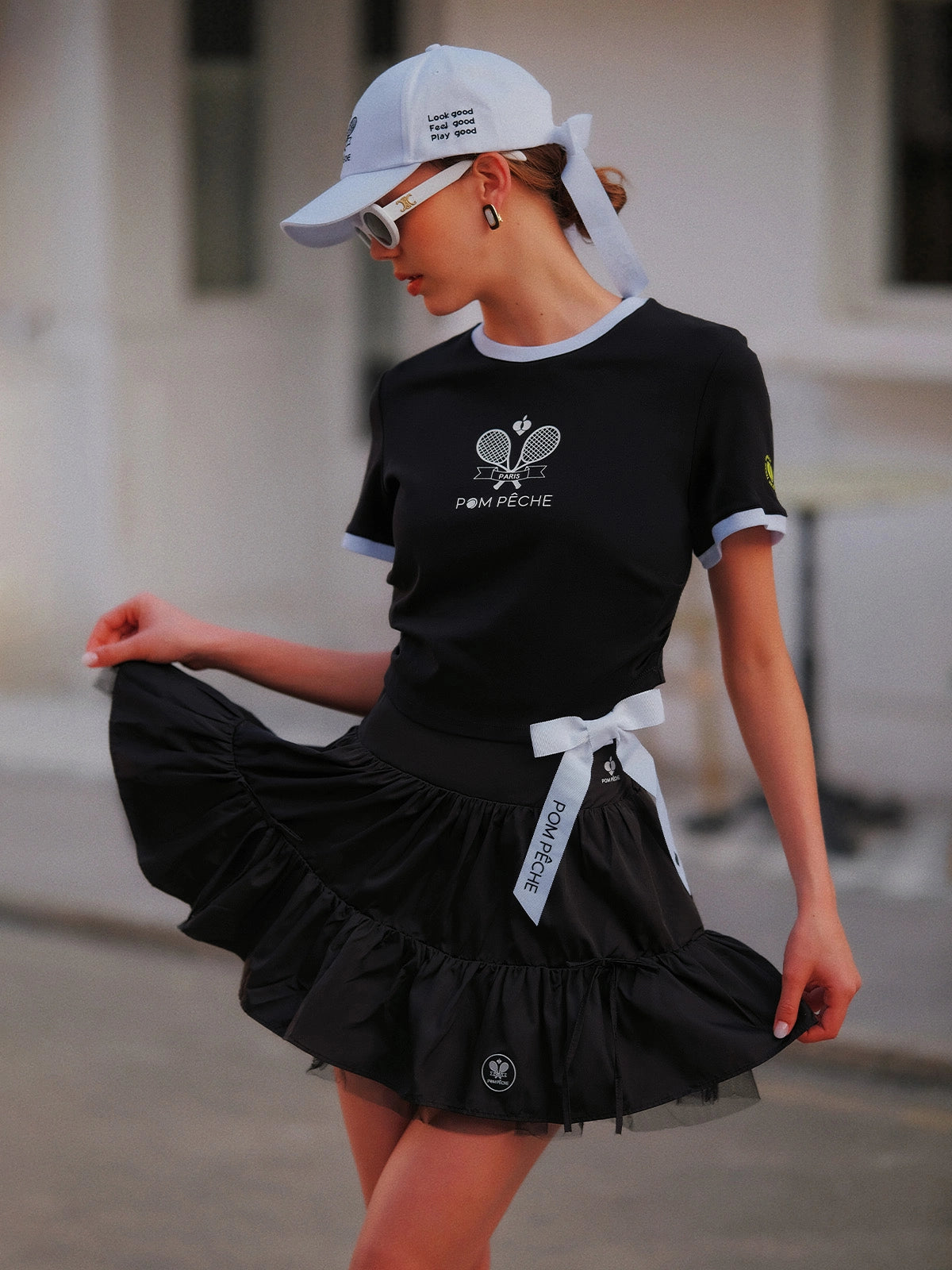 Contrast Trim Tennis Tee black, dynamic street editorial shot with flowing black ruffle skirt and Pom Pêche white bow, Pom Pêche cap