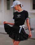 Contrast Trim Tennis Tee black, dynamic street editorial shot with flowing black ruffle skirt and Pom Pêche white bow, Pom Pêche cap