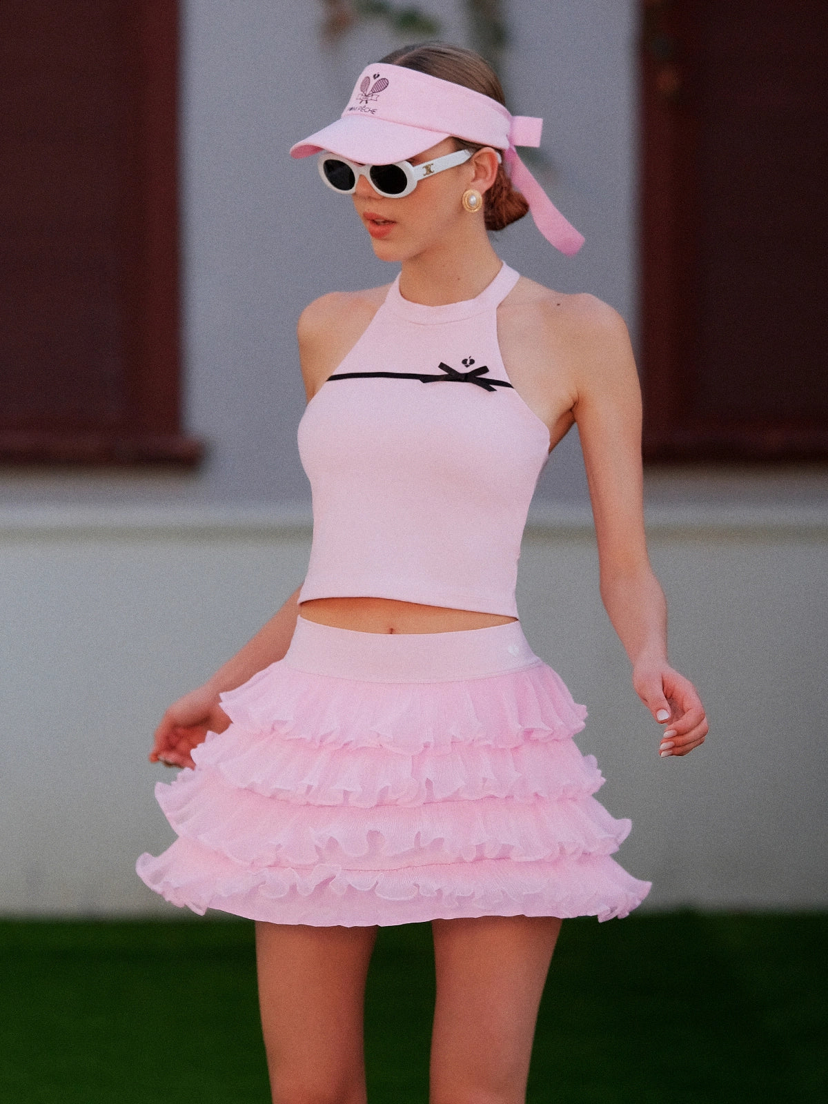 Pink Bow Halter Tennis Top worn with pink ruffle tennis skirt and Pom Pêche visor, full court-ready outfit on grass court