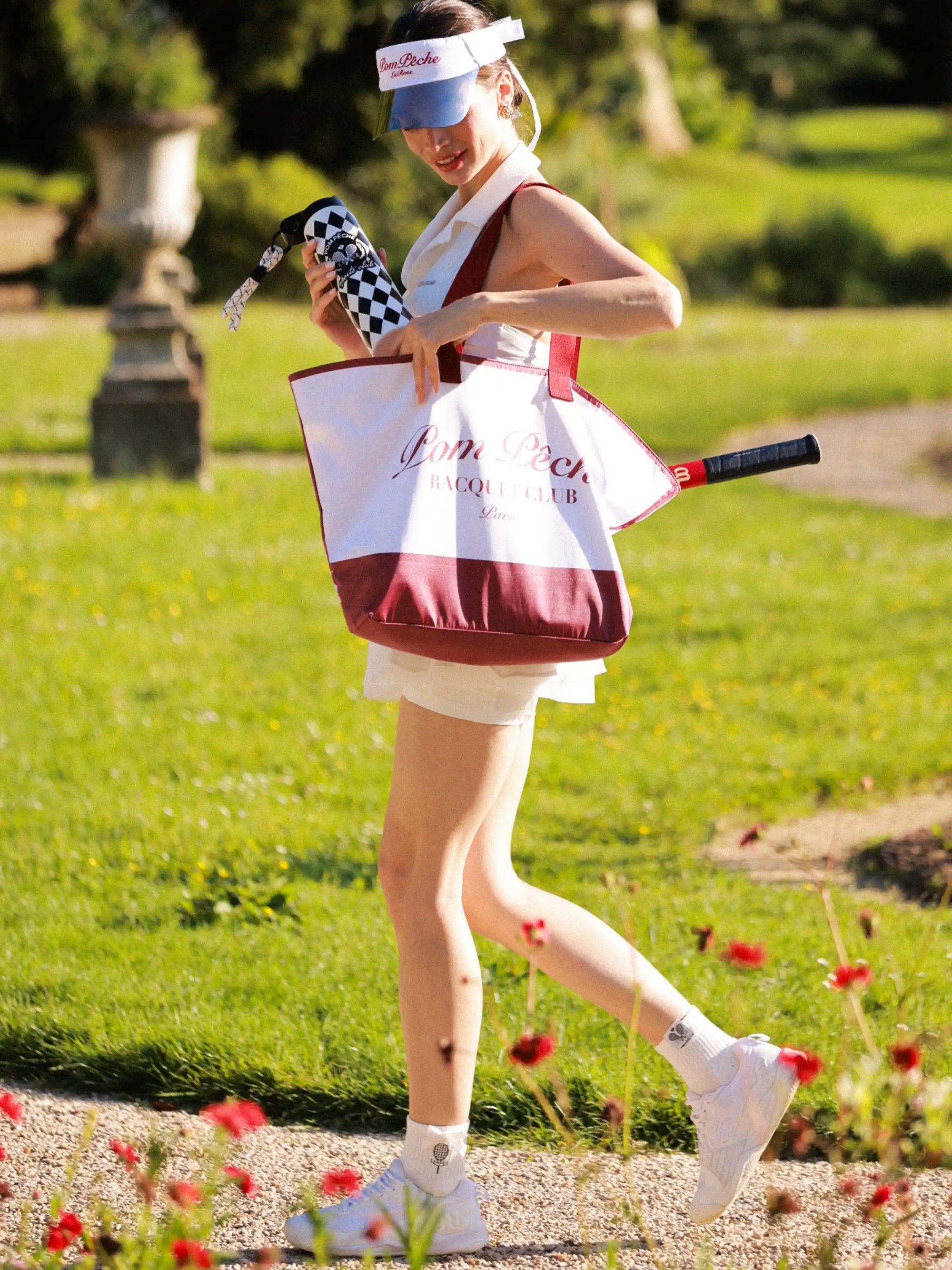 Pom Pêche 750ml Insulated Tumbler in black and white harlequin – model walking in Paris garden holding tumbler and Pom Pêche Racquet Club canvas tote bag with tennis racket, styled with white Poise polo halter top, Pom Pêche La Rose mirror visor, white tennis sneakers and Pom Pêche rose embroidered socks