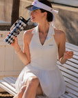 Lifestyle shot of Pom Pêche 750ml Insulated Tumbler in black and white harlequin – model drinking through straw lid on Paris garden white bench, Pom Pêche Sports Club crest visible, styled with white Poise polo halter top, Pom Pêche La Rose mirror visor, gold earrings and pearl bracelet