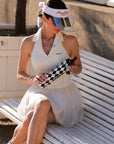 Close-up of Pom Pêche 750ml Insulated Tumbler in black and white harlequin – model opening black straw lid showing Pom Pêche Sports Club Paris crest badge and braided carry strap, Paris garden white bench background, Pom Pêche La Rose mirror visor and gold earrings