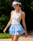 Full body view of Pom Pêche x Betty Boop Poise™ Polo Tank in white – polo collar, vertical Pom FlexRib™ ribbed texture with Betty Boop circular badge and full-zip front, styled with sky blue Betty Boop Cake Skirt, blue Pom Pêche visor and white bow ribbon walking on Paris garden path