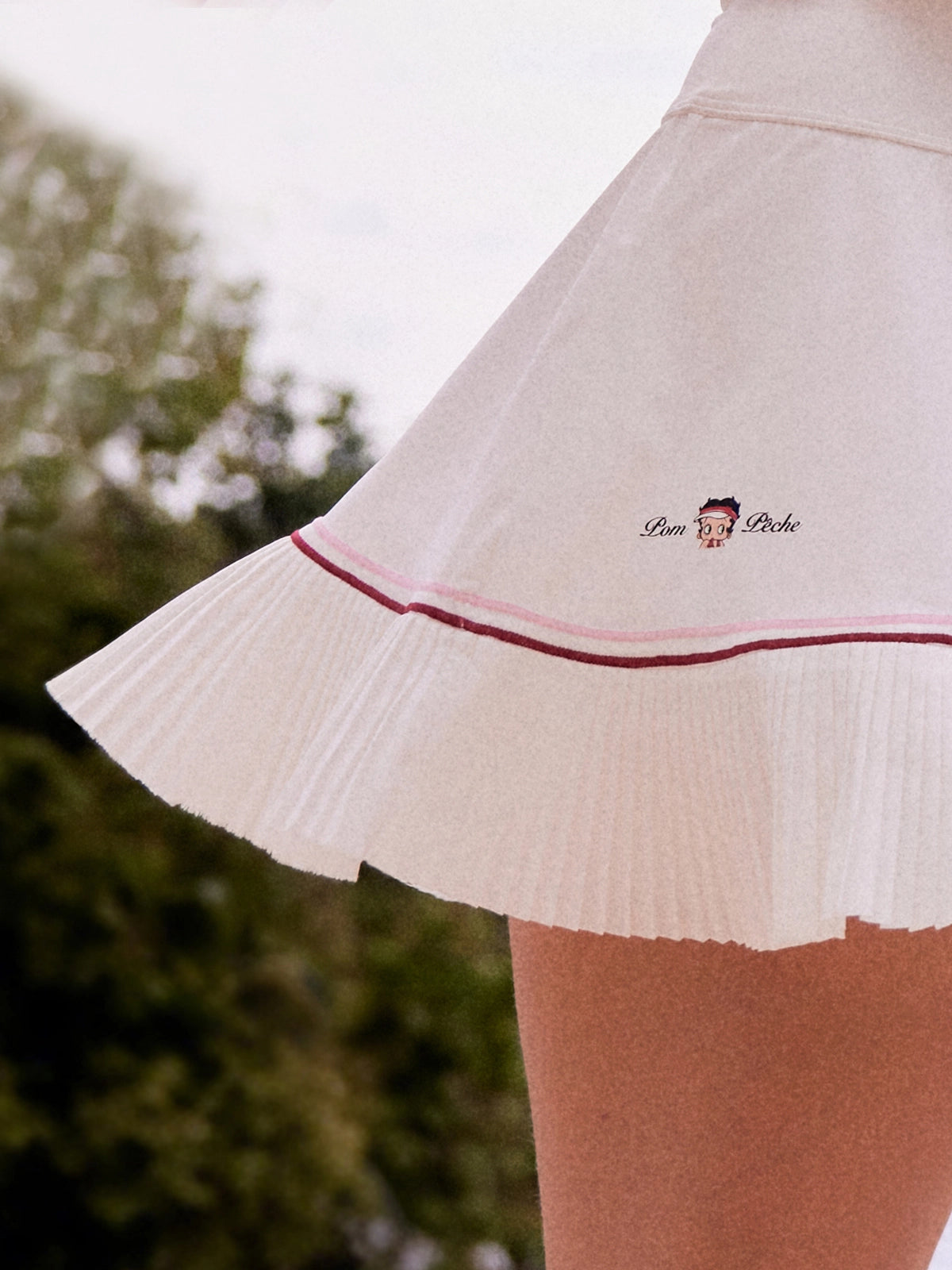 Macro detail of Pom Pêche x Betty Boop white Pleated Panel Skirt showing asymmetric heat-set pleated hem with pink and burgundy contrast double piping and Betty Boop Pom Pêche embroidered logo