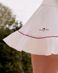 Macro detail of Pom Pêche x Betty Boop white Pleated Panel Skirt showing asymmetric heat-set pleated hem with pink and burgundy contrast double piping and Betty Boop Pom Pêche embroidered logo