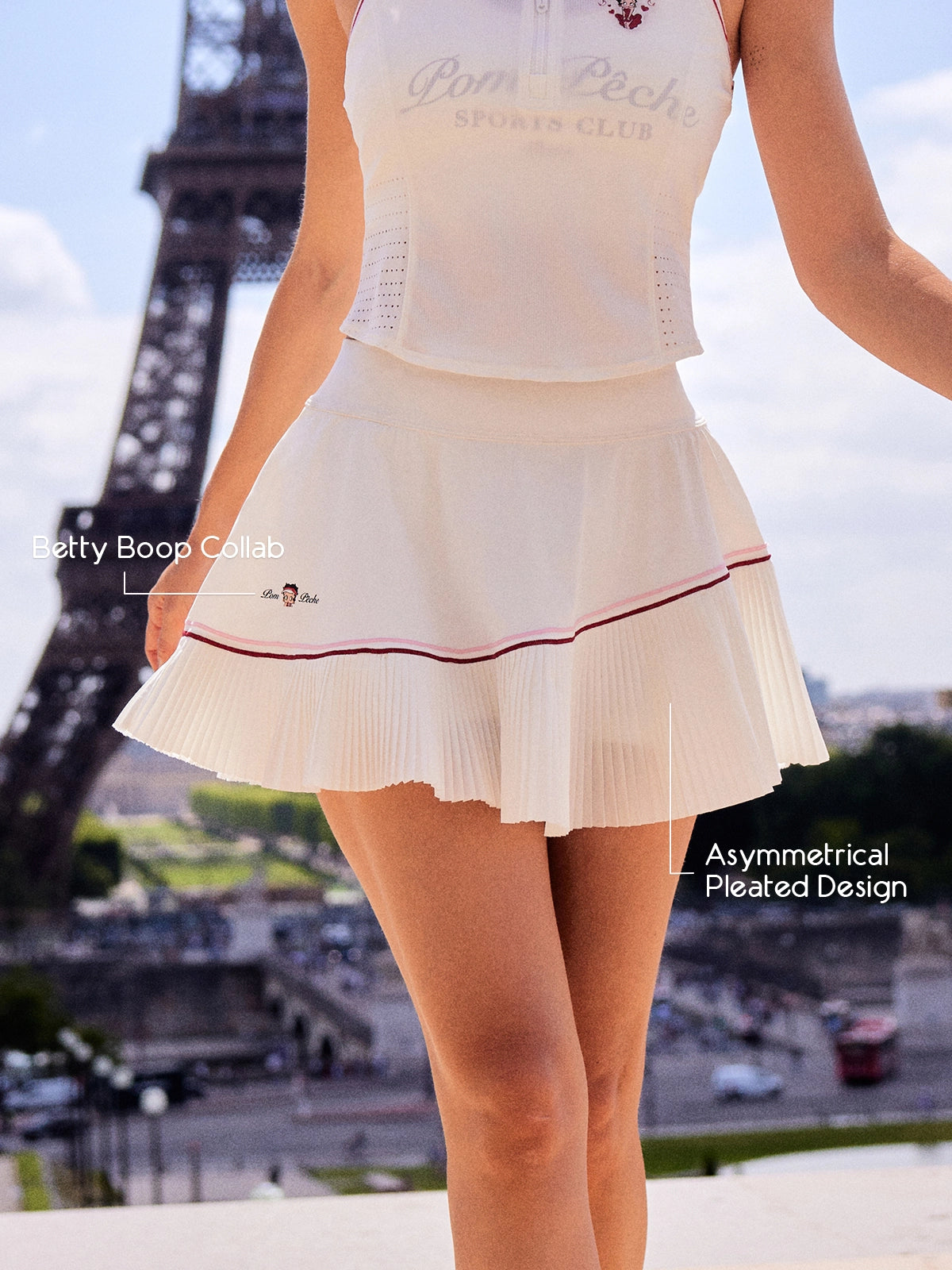 Pom Pêche x Betty Boop white Pleated Panel Skirt feature callout – Betty Boop Collab embroidery and Asymmetrical Pleated Design annotated, styled with matching white polo halter top, Eiffel Tower Paris