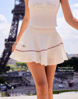 Pom Pêche x Betty Boop white Pleated Panel Skirt feature callout – Betty Boop Collab embroidery and Asymmetrical Pleated Design annotated, styled with matching white polo halter top, Eiffel Tower Paris
