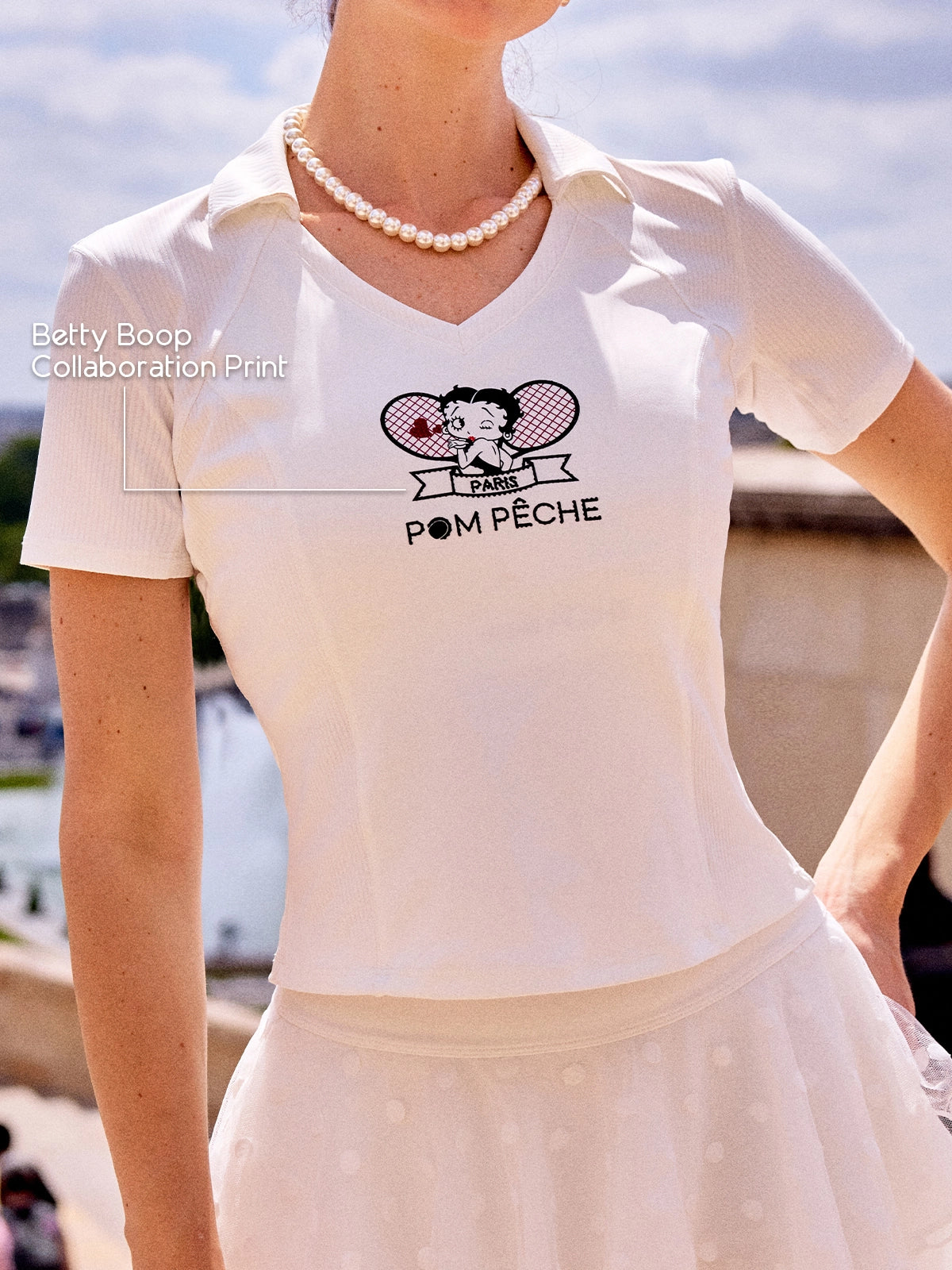 Close-up of Pom Pêche x Betty Boop white Tennis Tee with Betty Boop Collaboration Print callout – Betty Boop Paris twin-racket graphic with Pom Pêche script, V-neck polo collar, styled with pearl necklace on Paris Seine riverbank