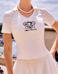 Close-up of Pom Pêche x Betty Boop white Tennis Tee with Betty Boop Collaboration Print callout – Betty Boop Paris twin-racket graphic with Pom Pêche script, V-neck polo collar, styled with pearl necklace on Paris Seine riverbank