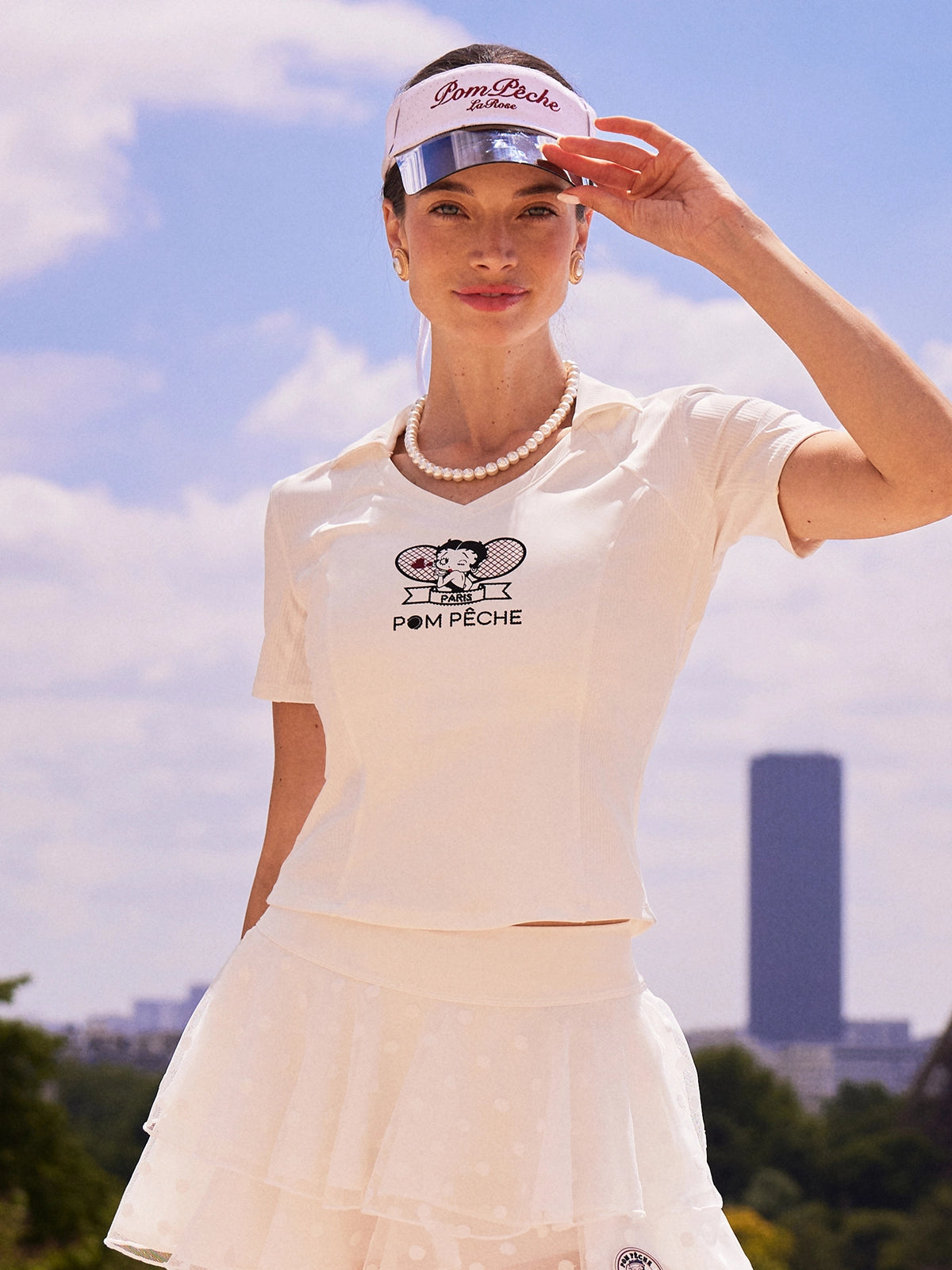 Pom Pêche x Betty Boop Tennis Tee in white – V-neck polo collar with Betty Boop Paris twin-racket graphic and Pom Pêche script, slim-fit short sleeve silhouette, styled with white Betty Boop Cake Skirt, Pom Pêche La Rose mirror visor and pearl necklace against Paris blue sky