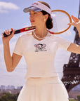 Pom Pêche x Betty Boop white Tennis Tee with UPF 50+ and Waist Accentuating Design callouts – model holding vintage wooden tennis racket, Betty Boop Paris graphic, styled with white Cake Skirt, Pom Pêche mirror visor and pearl necklace in front of Eiffel Tower