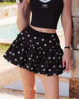 Full body view of Pom Pêche Bubble A-Line Tennis Skirt in black – high-waisted band with Pom Pêche logo, French bow side detail and voluminous A-line hem, styled with black tank top poolside
