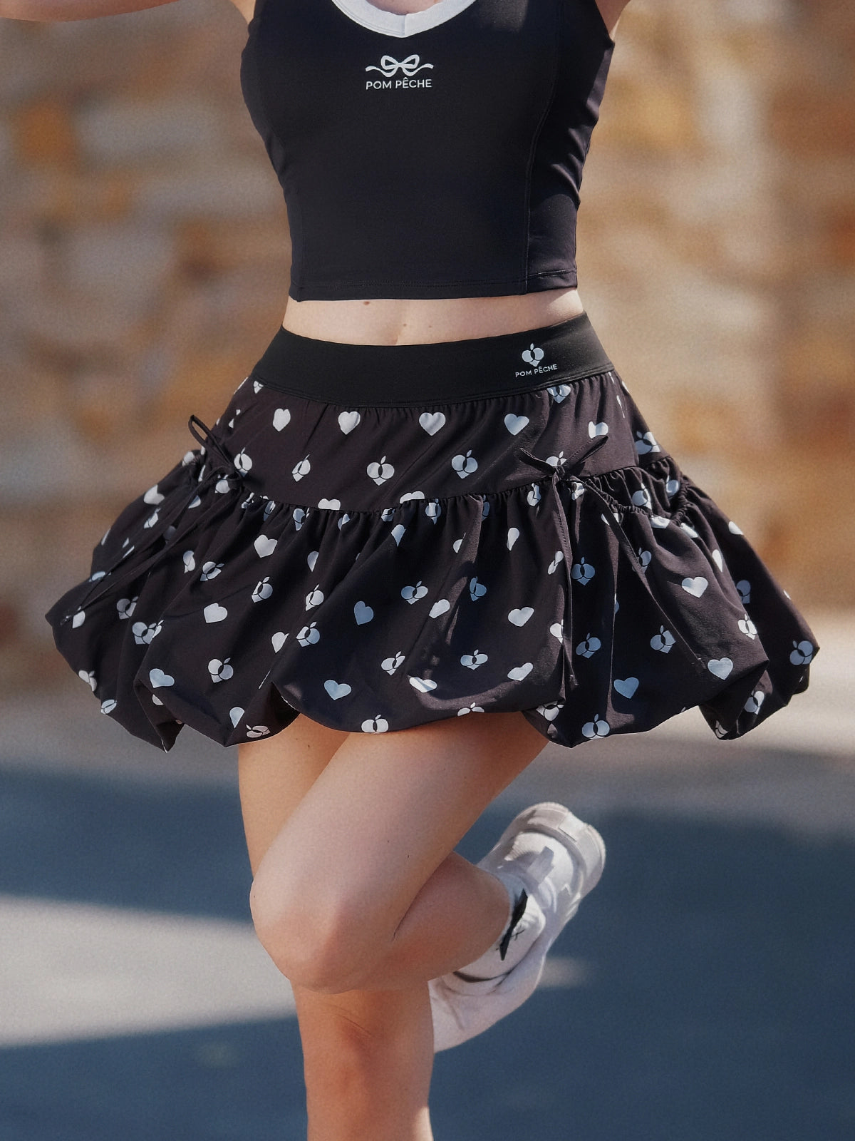 Pom Pêche Bubble A-Line Tennis Skirt in black – voluminous bubble hem with white heart and Pom Pêche logo print flaring during jump, styled with black Pom Pêche tank top